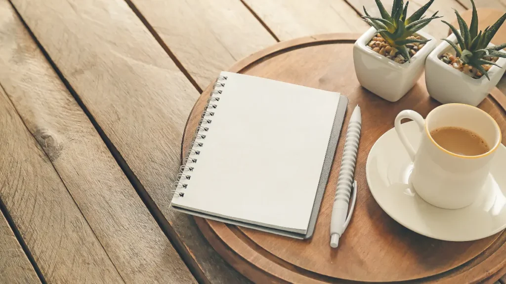 Minimalist wooden desk with a blank open notebook, a silver pen, a white cup of coffee, and small potted succulent plants in soft morning light